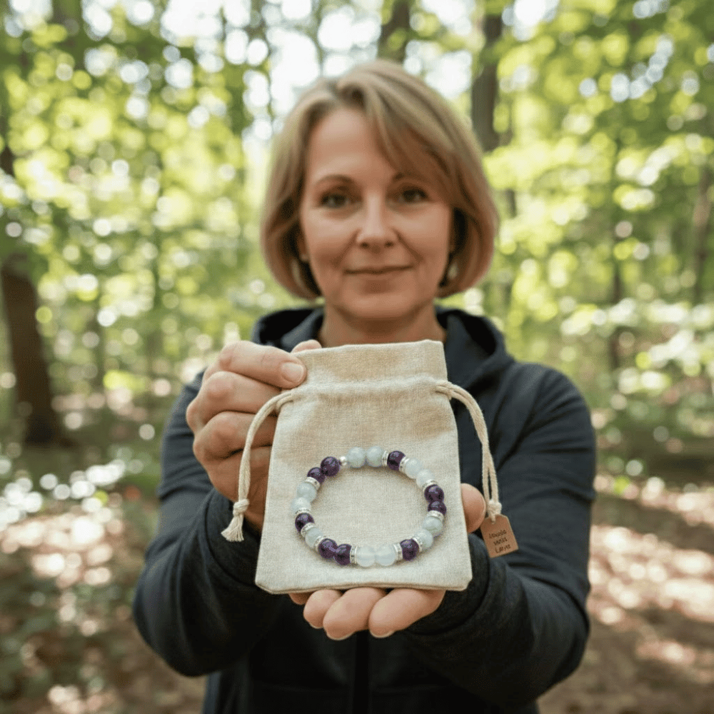 Una mujer en un bosque soleado ofrece una Pulsera Serena - Alivio Natural para la Menopausia, mostrando sus piedras blancas y moradas para la menopausia de una bolsa con cordón y sonriendo amablemente mientras comparte alivio síntomas menopausia.