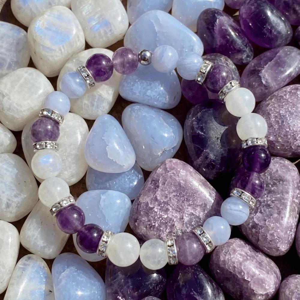 Pulsera Serena - Alivio Natural para la Menopausia, con cuentas moradas, blancas y translúcidas, separadores plateados y exhibida sobre piedras en tonos púrpura, azul y blanco para aliviar los síntomas de la menopausia.