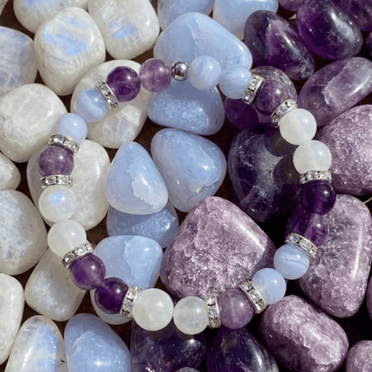 Pulsera Serena - Alivio Natural para la Menopausia, con cuentas moradas, blancas y translúcidas, separadores plateados y exhibida sobre piedras en tonos púrpura, azul y blanco para aliviar los síntomas de la menopausia.